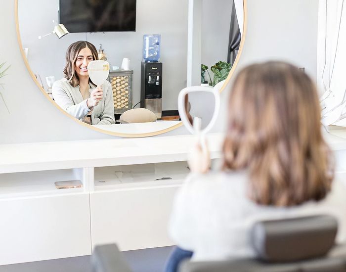 Woman smiling in salon mirror with handheld mirror.