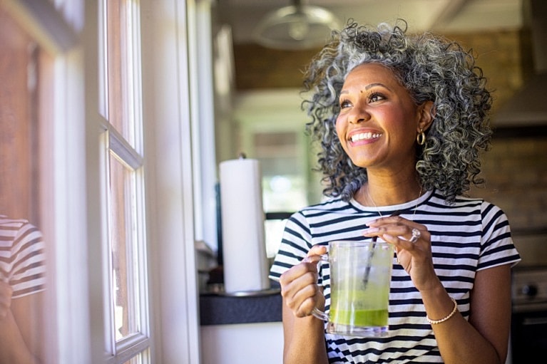 Smiling woman holding a green drink by window.